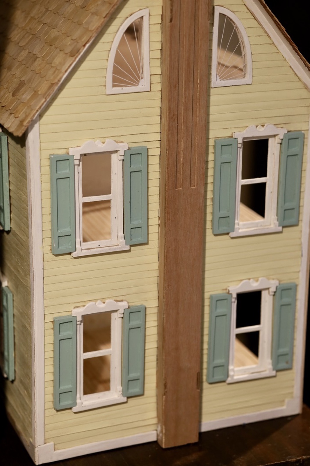 Front exterior of the handmade doll house with pale yellow siding, green shutters, and wood roof shingles